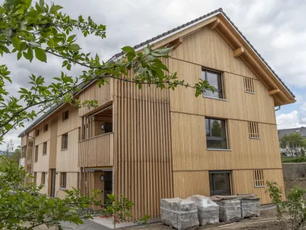 Aussenansicht eines neu errichteten Mehrfamilienhauses in moderner Holzbauweise mit senkrechter Holzfassade und Satteldach. Balkone und Fassadenelemente sind aus Holz gestaltet. Im Vordergrund sind Pflanzen und Baustellenmaterialien zu sehen, im Hintergrund ein Wohnquartier unter bewölktem Himmel.