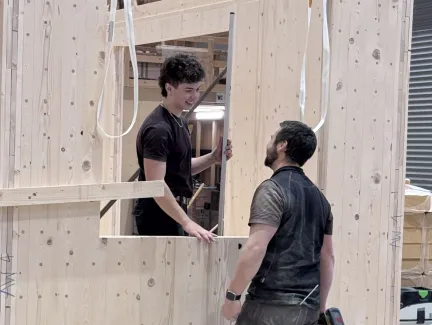 Zwei Mitarbeitende der Zimmerei Hirschi montieren in der Werkhalle ein vorgefertigtes Holzelement. Sie arbeiten an einer massiven Holzrahmenkonstruktion mit Fensteröffnung und stimmen sich dabei direkt vor Ort ab.