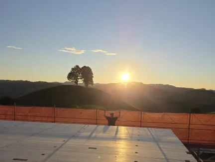 Blick über eine neu verlegte Holzdecke auf einer Baustelle bei Sonnenuntergang. Im Hintergrund steht ein Zimmermann am Gerüst vor der hügeligen Landschaft und hebt beide Arme in die Höhe, während die tief stehende Sonne über den Hügeln scheint.