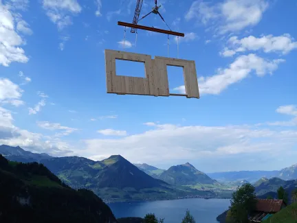 Vorgefertigtes Holzwandelement mit Fenster- und Türöffnung wird von einem Kran über einer alpinen Berglandschaft transportiert. Im Hintergrund sind grüne Berghänge und ein See zu sehen, welche die präzise Montage von Holzbauelementen in anspruchsvollem Gelände veranschaulichen.