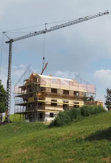Ein mehrstöckiges Holzhaus im Bau, umgeben von Gerüsten und einem Baukran, steht auf einer grünen Anhöhe unter blauem Himmel mit einigen Wolken.
