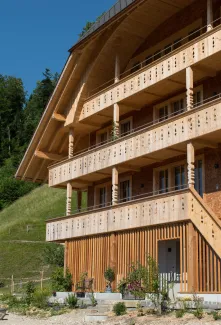 Mehrstöckiges Holzhaus im traditionellen Schweizer Stil mit kunstvoll verzierten Holzbalkonen und grosszügigem Dachüberstand. Die Fassade besteht aus hellem, massivem Holz, umgeben von einer grünen Hanglandschaft bei klarem Himmel.
