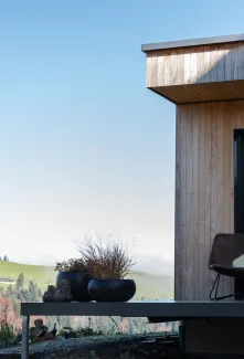 Aussenansicht eines modernen Holzhauses mit klarer Linienführung. Auf der erhöhten Terrasse stehen zwei Stühle mit Kissen und ein kleiner Tisch vor einer grossen Glasfront. Die Fassade besteht aus senkrechter Holzschalung, im Hintergrund sind Hügel, Bäume und eine ländliche Landschaft bei klarem Himmel zu sehen.