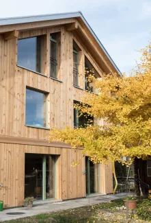 Aussenansicht eines mehrgeschossigen Wohnhauses in moderner Holzbauweise mit senkrechter Holzfassade und grossen Fenstern. Vor dem Gebäude steht ein Baum mit gelben Herbstblättern, der Teile der Fassade überragt. Der Eingangsbereich und der Garten mit Pflanzen und Pflasterflächen sind im Vordergrund sichtbar, bei hellem Tageslicht.