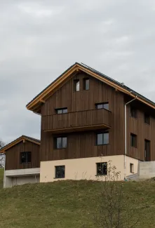 Neubau eines modernen Holzhauses am Hang mit dunkler Holzfassade, Balkon und grosser Fensterfront. Das Gebäude steht in ländlicher Umgebung mit Wiese und weiteren Häusern im Hintergrund unter bewölktem Himmel.