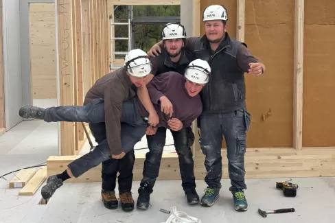 Vier Zimmerleute posieren lachend auf der Baustelle eines Holzhauses. Die fröhliche Stimmung zeigt den Teamgeist und die Freude am Handwerk bei der Zimmerei Hirschi AG.