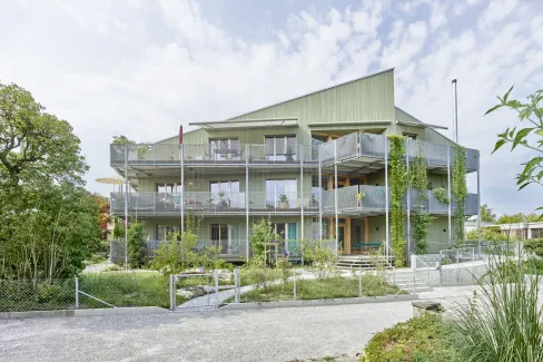 Aussenansicht eines modernen, mehrstöckigen Wohngebäudes in Holzbauweise mit hellgrüner Fassade. Das Haus verfügt über grosse Balkone mit Metallgeländern und Kletterpflanzen, die vertikal an den Fassadenelementen emporwachsen. Vor dem Gebäude liegt ein gepflegter Garten mit jungen Bäumen, Sträuchern und geschwungenen Wegen. Die Architektur wirkt offen und einladend, mit vielen Fenstern und einer klar strukturierten Formensprache. Im Vordergrund sind ein Kiesweg und ein Zaun sichtbar, während der Himmel leic