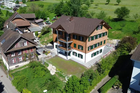 Luftaufnahme eines frisch renovierten, mehrstöckigen Holzhauses im traditionellen Stil mit brauner Schindelfassade und grünen Fensterläden. Das Gebäude steht inmitten einer ländlichen Umgebung mit Wiesen, Bäumen und benachbarten Wohn- und Wirtschaftsbauten. Vor dem Haus befindet sich ein neu angelegter Vorplatz, während rundherum noch Bereiche der Gartengestaltung und Bauarbeiten sichtbar sind. Im Hintergrund erstreckt sich eine grüne Hügellandschaft mit verstreuten Obstbäumen und einigen modernen Wohnhäuse