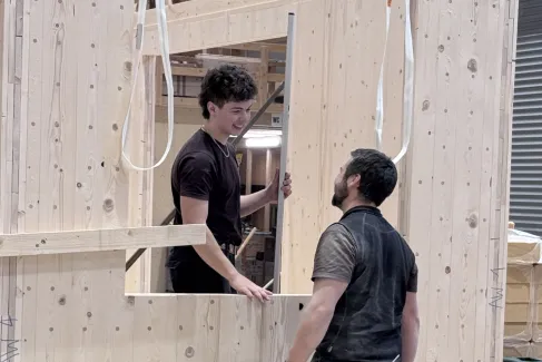 Zwei Mitarbeitende der Zimmerei Hirschi montieren in der Werkhalle ein vorgefertigtes Holzelement. Sie arbeiten an einer massiven Holzrahmenkonstruktion mit Fensteröffnung und stimmen sich dabei direkt vor Ort ab.