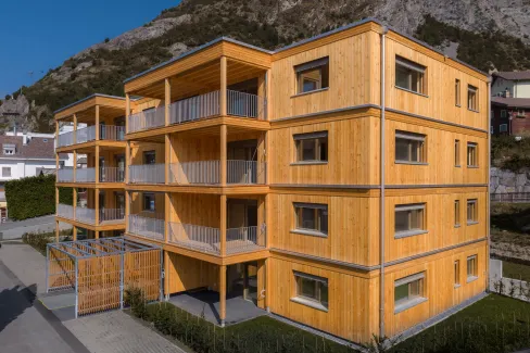 Aussenansicht eines modernen, viergeschossigen Mehrfamilienhauses in Holzbauweise vor einer felsigen Bergkulisse. Das Gebäude verfügt über eine vertikale Holzfassade sowie grosszügige, überdachte Balkone mit sichtbarer Holzkonstruktion und Metallgeländern.