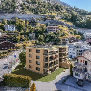 Visualisierung eines modernen Mehrfamilienhauses in Holzbauweise mit grossen Balkonen und begrüntem Dach mit Solaranlage. Das Gebäude fügt sich in eine alpine Dorflandschaft mit traditionellen Häusern, Strassen und einem bewaldeten Berghang im Hintergrund ein