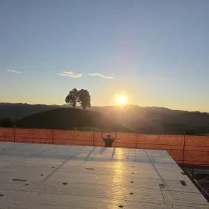 Blick über eine neu verlegte Holzdecke auf einer Baustelle bei Sonnenuntergang. Im Hintergrund steht ein Zimmermann am Gerüst vor der hügeligen Landschaft und hebt beide Arme in die Höhe, während die tief stehende Sonne über den Hügeln scheint.