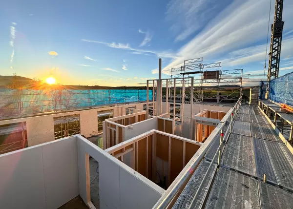 Baustelle eines Holzbaus bei Sonnenuntergang. Die beleuchtete Holzkonstruktion und das eingerüstete Gebäude zeigen den fortgeschrittenen Aufbau eines modernen Holzhauses durch die Zimmerei Hirschi AG.