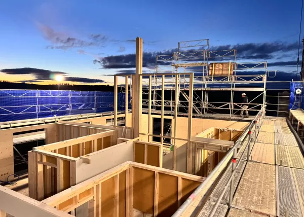 Baustelle eines Holzbaus bei Sonnenuntergang. Die beleuchtete Holzkonstruktion und das eingerüstete Gebäude zeigen den fortgeschrittenen Aufbau eines modernen Holzhauses durch die Zimmerei Hirschi AG.