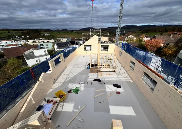 Baustelle eines Holzbaus bei Sonnenuntergang. Die beleuchtete Holzkonstruktion und das eingerüstete Gebäude zeigen den fortgeschrittenen Aufbau eines modernen Holzhauses durch die Zimmerei Hirschi AG.
