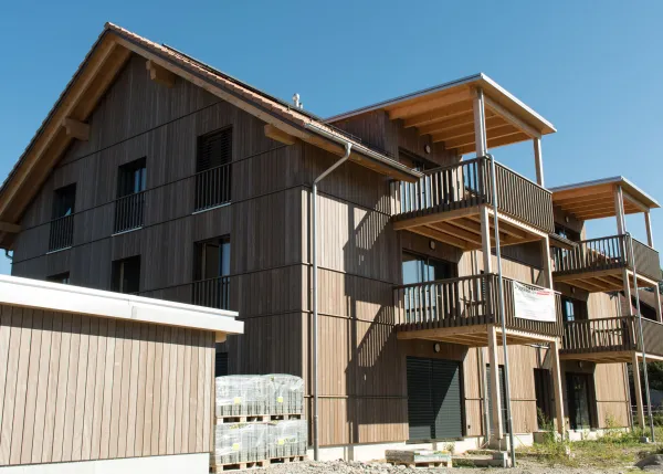 Aussenansicht eines mehrgeschossigen Wohnhauses in Holzbauweise mit Satteldach und integrierten Solarpanels. Die Fassade besteht aus senkrechter Holzschalung, die Balkone sind aus Holz gefertigt. Das Gebäude steht in einem Wohnquartier mit Hecken und Nachbarbauten, im Vordergrund verlaufen Bahnoberleitungen vor blauem Himmel.