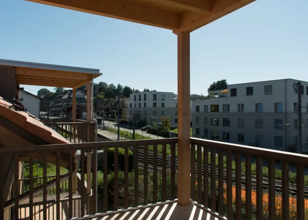 Aussenansicht eines mehrgeschossigen Wohnhauses in Holzbauweise mit Satteldach und integrierten Solarpanels. Die Fassade besteht aus senkrechter Holzschalung, die Balkone sind aus Holz gefertigt. Das Gebäude steht in einem Wohnquartier mit Hecken und Nachbarbauten, im Vordergrund verlaufen Bahnoberleitungen vor blauem Himmel.