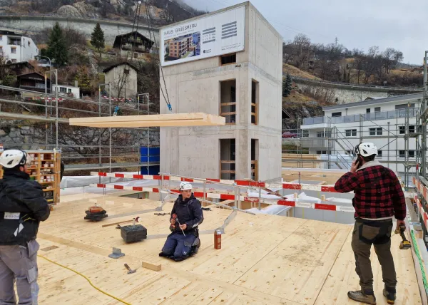 Zimmerleute arbeiten an der Holzrahmenkonstruktion eines mehrgeschossigen Gebäudes auf einer Baustelle. Die tragenden Wände aus Holz werden vor Ort montiert, während ein Baukran über dem Bauplatz steht. Im Hintergrund erstrecken sich Dorfhäuser und eine beeindruckende alpine Berglandschaft
