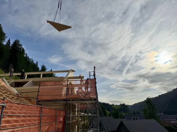 Zimmerleute montieren während der Aufrichtearbeiten eine vorgefertigte Holzkonstruktion auf einem Gebäude. Ein Kran hebt ein grosses Dachelement an seinen Platz, während mehrere Fachkräfte auf dem Dach die Montage begleiten.