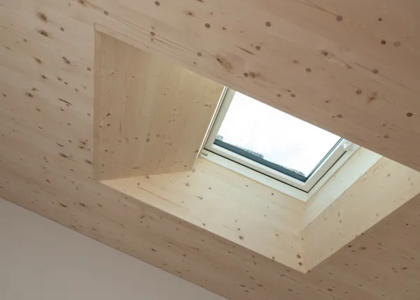 Modernes Badezimmer mit grossformatigen grauen Fliesen, Badewanne und Waschtisch mit hölzernem Unterschrank. Die helle, geneigte Holzdecke und die sichtbaren Holzelemente schaffen einen warmen Kontrast zur schlichten Badezimmereinrichtung.